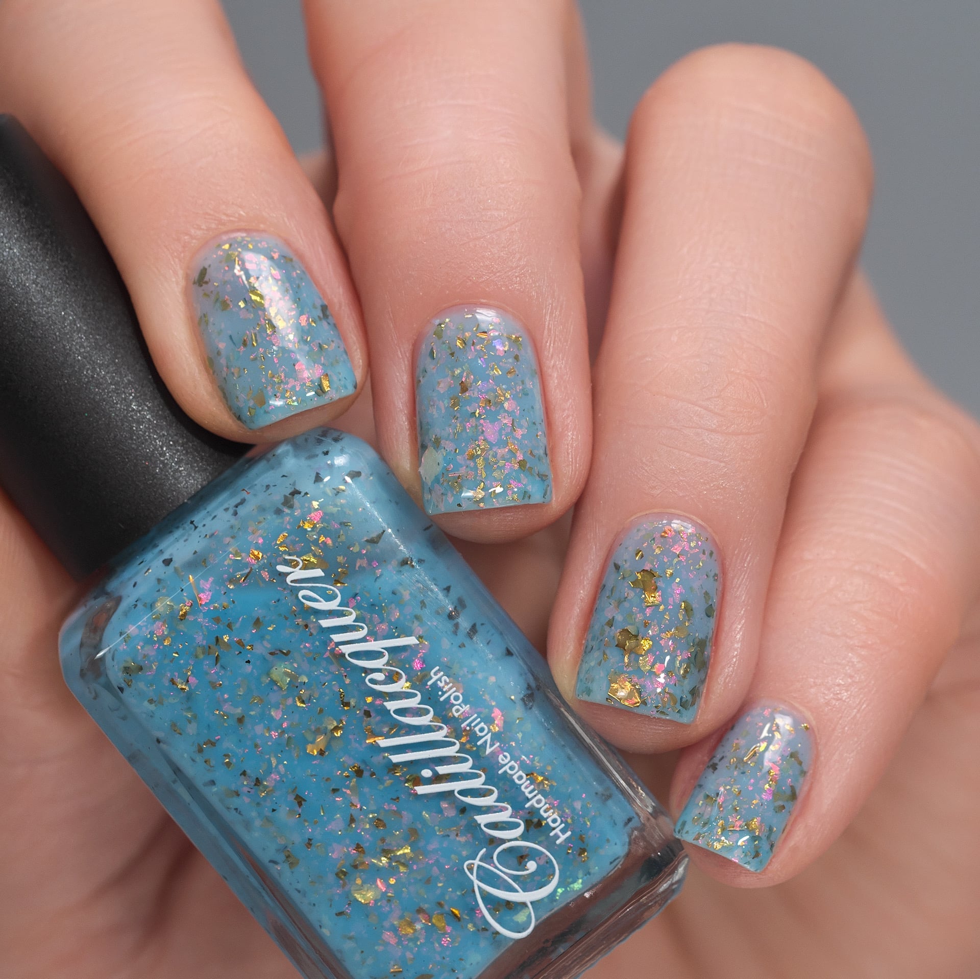 Cadillacquer Slow Mornings light blue gold chameleon iridescent crelly nail polish, bottle held with four-nail swatch, light skin, studio lighting