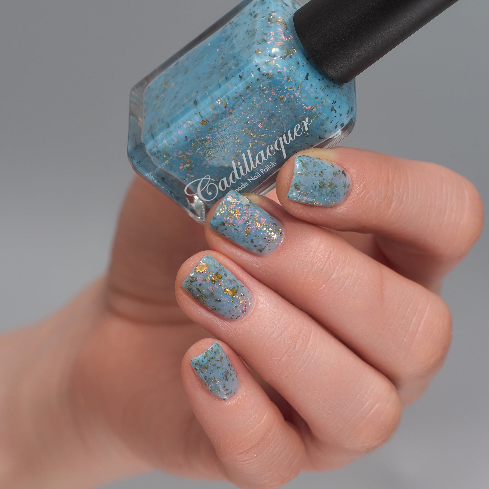 Cadillacquer Slow Mornings light blue gold chameleon iridescent crelly nail polish, bottle held with four-nail swatch, light skin, studio lighting
