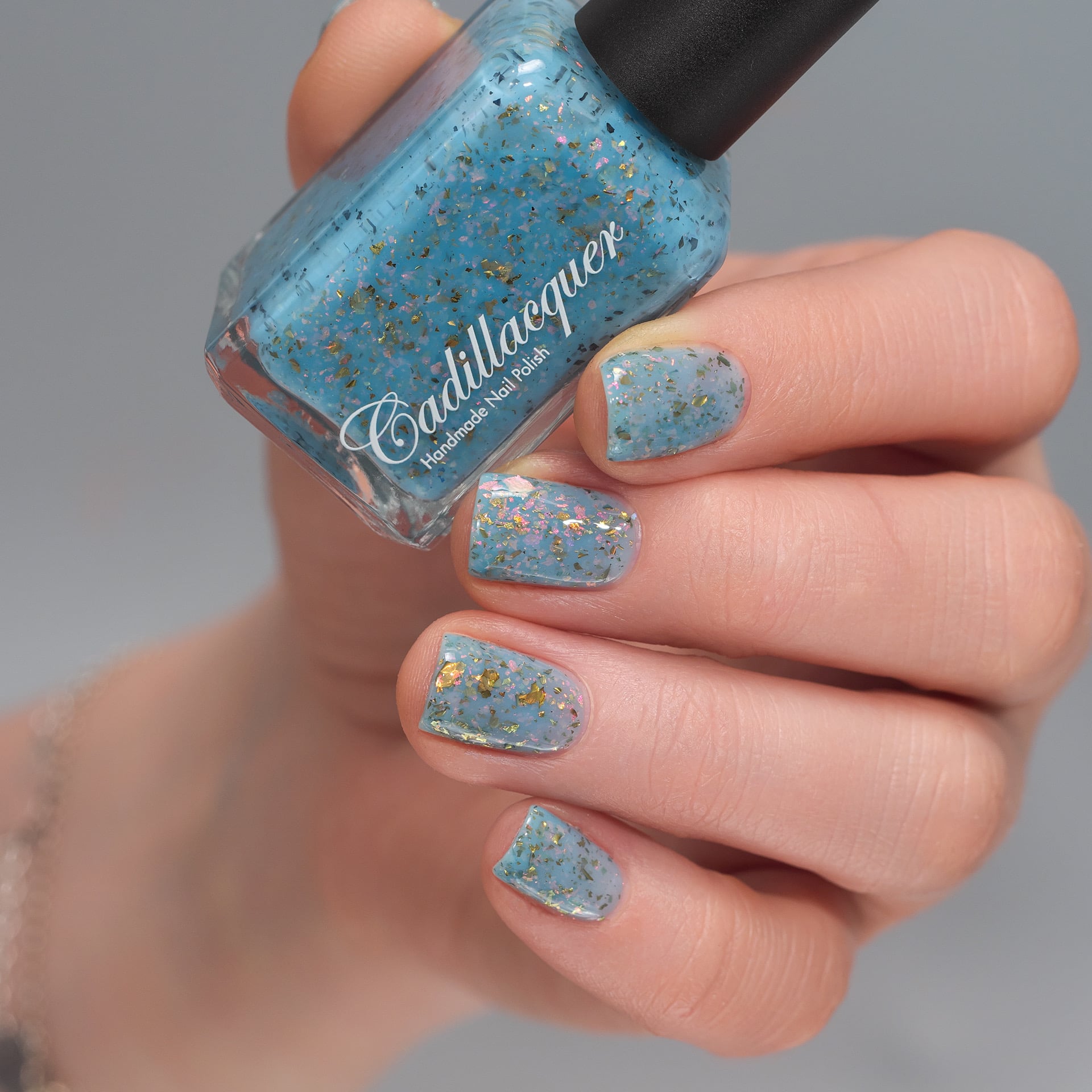 Cadillacquer Slow Mornings light blue gold chameleon iridescent crelly nail polish, bottle held with four-nail swatch, light skin, studio lighting