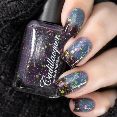 Thermal glitter and flake Cadillacquer nail polish in Strange Happenings, shown in bottle held in hand alongside five swatched nails demonstrating the cold blackened eggplant to warm dusty blue thermal transition, loaded with matte neon green glitter in multiple sizes and hot pink to orange iridescent flakes, light skin tone, studio lighting, swatched by @serpentine13