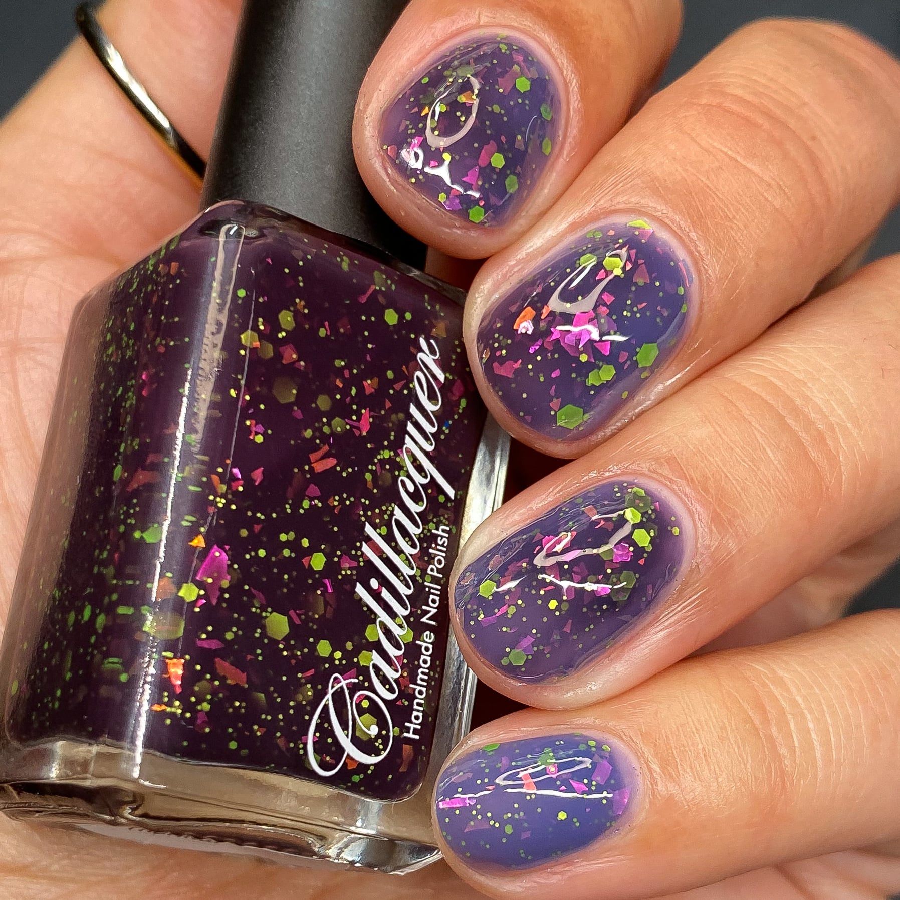 Thermal glitter-and-flake Cadillacquer nail polish in Strange Happenings, shown in bottle-in-hand with four swatched nails displaying the thermal transition from blackened eggplant (cold, ring finger) to dusty blue-violet (warm, index and middle), loaded with matte neon chartreuse-green glitter in multiple sizes and iridescent pink-to-orange flakes, light-medium skin tone, studio lighting, swatched by @heeymercedes