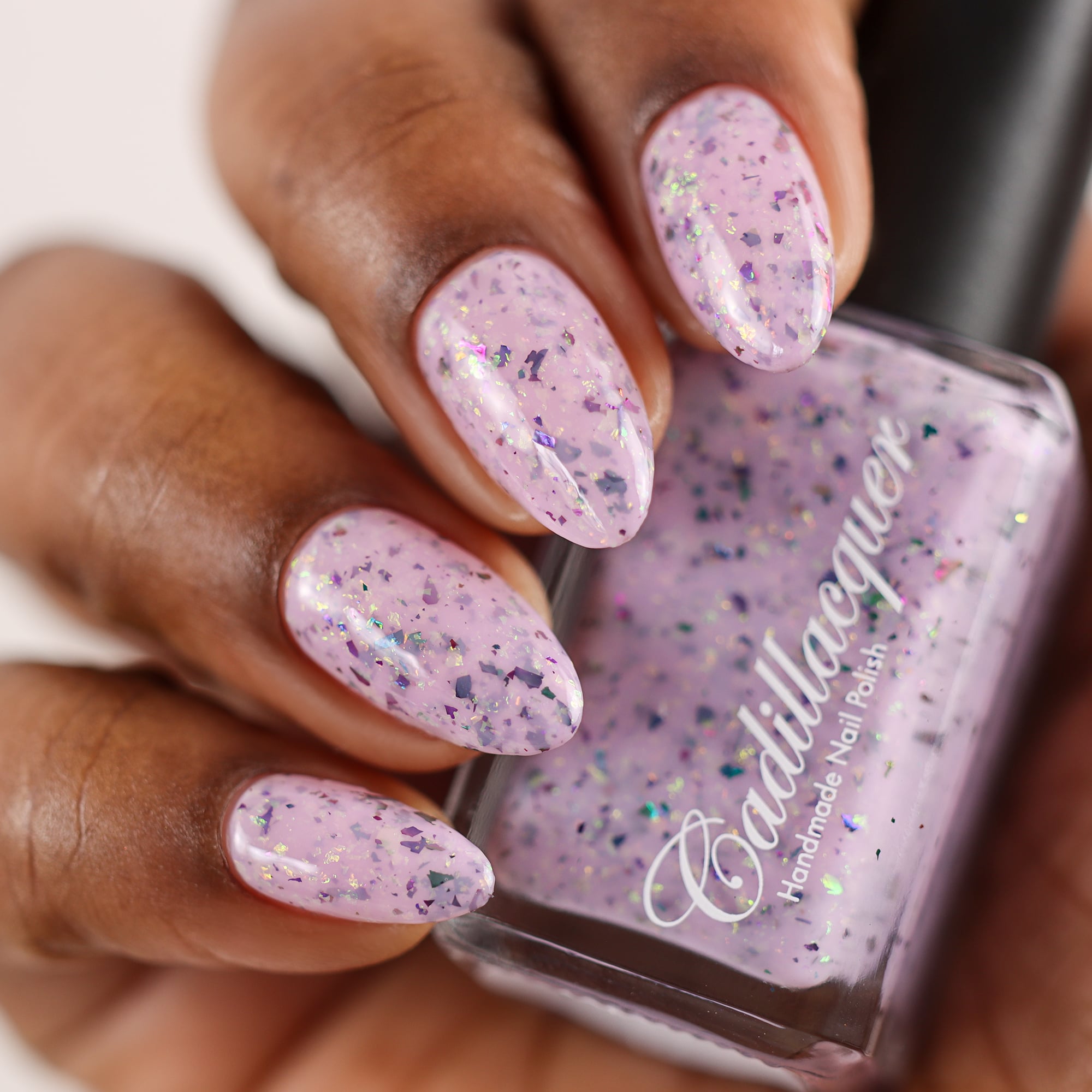 Cadillacquer Sweet Nothing pink teal chameleon iridescent crelly nail polish, bottle held with four-nail swatch, deep skin, studio lighting