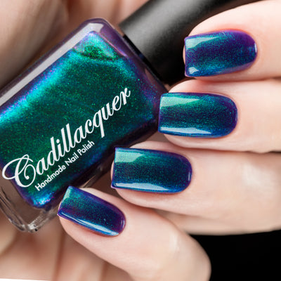 Multichrome holographic shimmer Cadillacquer nail polish in What Could Possibly Go Wrong?, shown as a bottle held in hand with four square nails swatched, shifting between deep indigo base, electric teal-green, vivid blue, and violet-purple with densely packed holographic green-to-blue shimmer particles, light skin tone, studio lighting, swatched by @rafinails