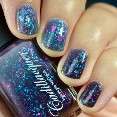Thermal glitter-and-flake Cadillacquer nail polish in What Lurks Within, shown on five short natural nails cycling between a dark blue-grey cold state and a lighter blue-grey warm state, densely packed with cyan-teal iridescent flakes that shift blue-violet and neon hot-pink matte glitter in mixed sizes, alongside the dark blackened-eggplant bottle, light skin tone, indoor warm lighting, swatched by @caitlinswatches