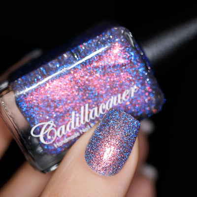 Multichrome flake and reflective glitter Cadillacquer nail polish in You Wished For This, shown as bottle held in hand with one swatched nail visible, bottle face dense with blue and rose-pink micro flakes flashing warm pink under direct flash, nail reads periwinkle-blue shifting to rose-pink at angle, light-medium skin tone, direct flash studio lighting, swatched by @yyulia_m