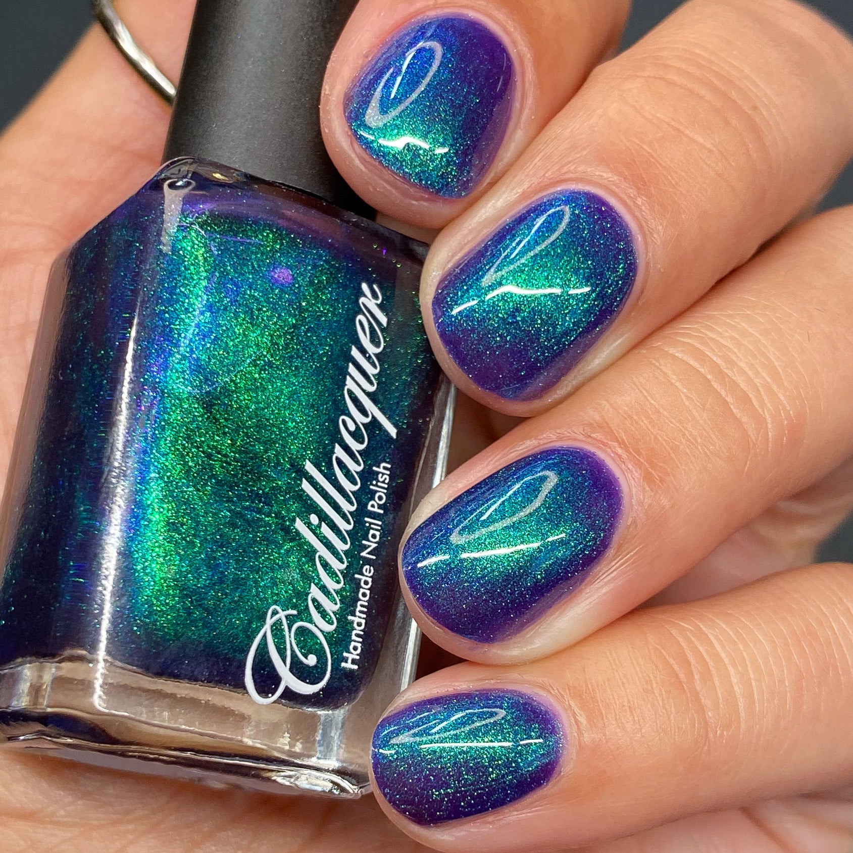 Multichrome shimmer Cadillacquer nail polish in What Could Possibly Go Wrong?, bottle held in hand alongside swatched nails showing deep violet-indigo base flashing vivid emerald green to blue-teal shimmer at different angles, light skin tone, studio lighting, swatched by @heeymercedes