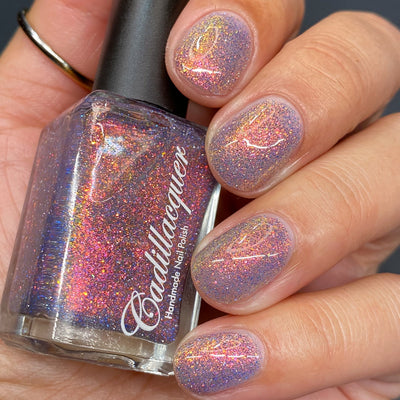 Multichrome flake and reflective glitter Cadillacquer nail polish in You Wished For This, shown in a bottle held in hand with five nails swatched, the sheer lavender-grey base packed with orange-to-pink-to-yellow iridescent micro flakes and scattered blue and charcoal reflective glitter particles, light-medium skin tone, studio lighting, swatched by @heeymercedes