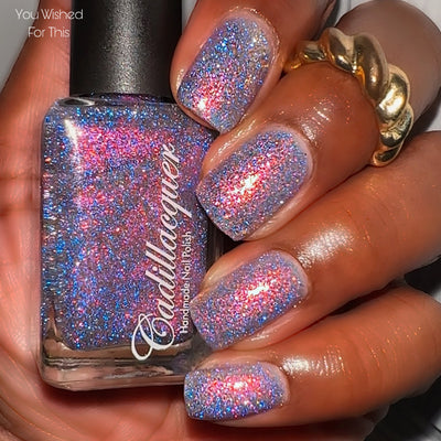 Flake and reflective glitter Cadillacquer nail polish in You Wished For This, shown in bottle held in hand with swatch on five square nails, sheer lavender-grey base packed with pink-to-orange iridescent micro flakes and electric blue reflective glitter that bursts with hot pink-red flash in direct light, medium-deep skin tone, direct flash lighting, swatched by @lacquer_is_life