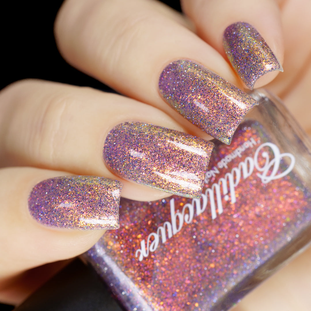 Holographic micro-flake and reflective glitter Cadillacquer nail polish in You Wished For This, shown as a swatch on four oval nails held around a Cadillacquer bottle, the nails read muted mauve-violet at rest and shift to warm copper-gold and burnt orange at angle, packed with fine red-orange-to-yellow iridescent micro flakes and scattered charcoal and blue holographic glitter, the bottle face reads deep copper-orange under direct studio light, light skin tone.