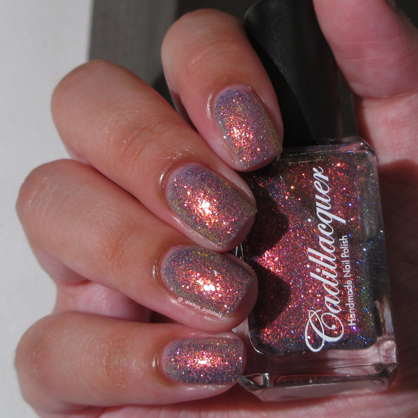 Multichrome flake and reflective glitter Cadillacquer nail polish in You Wished For This, shown on eight squoval nails with sheer mauve base packed with fine red-orange-to-yellow iridescent micro flakes and scattered blue and charcoal holographic glitter, bottle held in hand at right, light-medium skin tone, indoor warm lighting, swatched by @_always_polished_