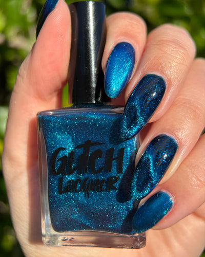 Heart of the Ocean by Glitch Lacquer, deep blue jelly with blue-to-purple magnetic pigment and blue glitters, bottle held in hand and swatched on five almond nails, light skin tone, bright daylight.