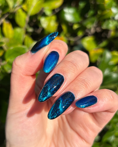 Heart of the Ocean by Glitch Lacquer on five almond nails outdoors against green foliage, deep blue magnetic jelly with blue glitters, light skin tone, bright sunlight.