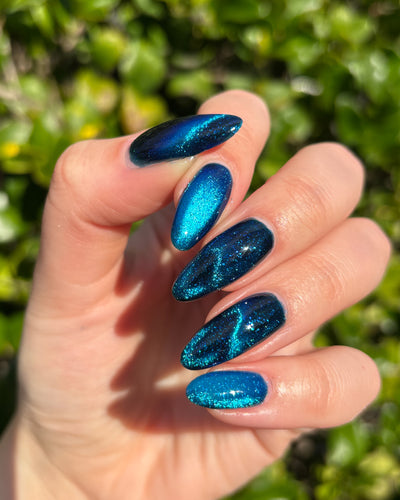 Heart of the Ocean by Glitch Lacquer on five almond nails showing magnetic cat-eye patterns, deep blue jelly base with blue glitters, light-medium skin tone, bright daylight.
