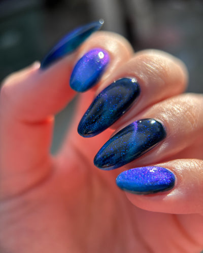 Heart of the Ocean by Glitch Lacquer on five almond nails, deep blue jelly with strong blue-to-purple magnetic stripe and blue glitters, light-medium skin tone, bright daylight.